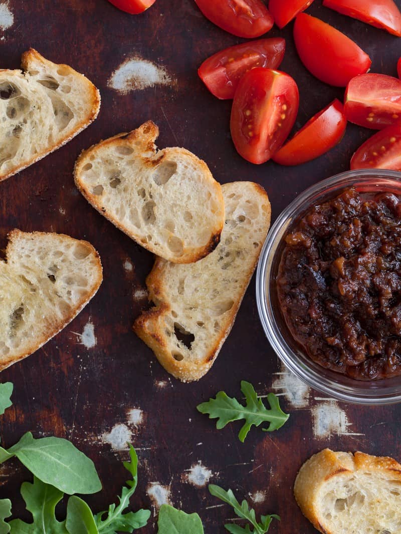 A close up of crostini with tomatoe wedges and a bowl of boozy bacon jam.