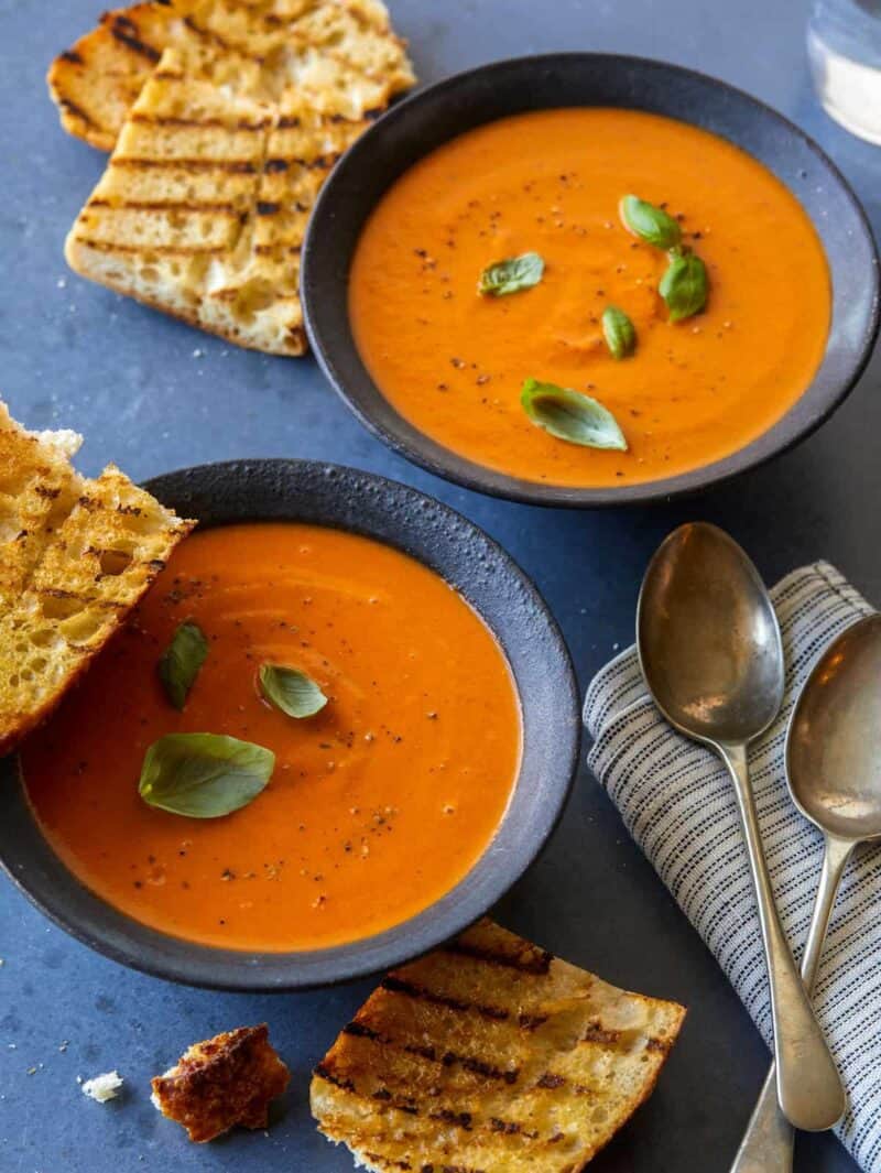 Two bowls of creamy roasted tomato basil soup with grilled bread, napkins, and spoons.