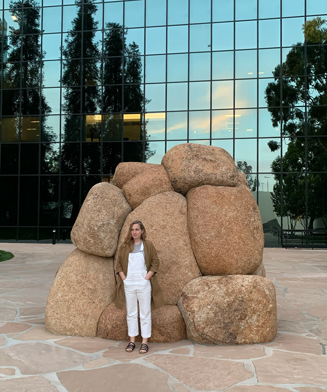 A relaxed woman standing in front of a stone sculpture with a building in the background.