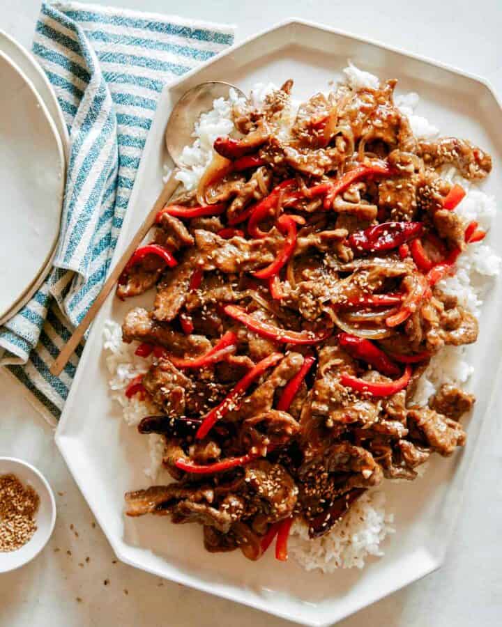 A platter of Szechuan Beef recipe with plates next to it and a ramekin of sesame seeds.