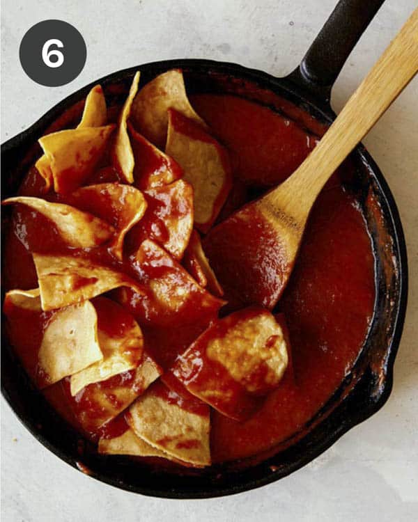 Tortilla chips in a skillet being gently folded together with a red sauce to make chilaquiles.