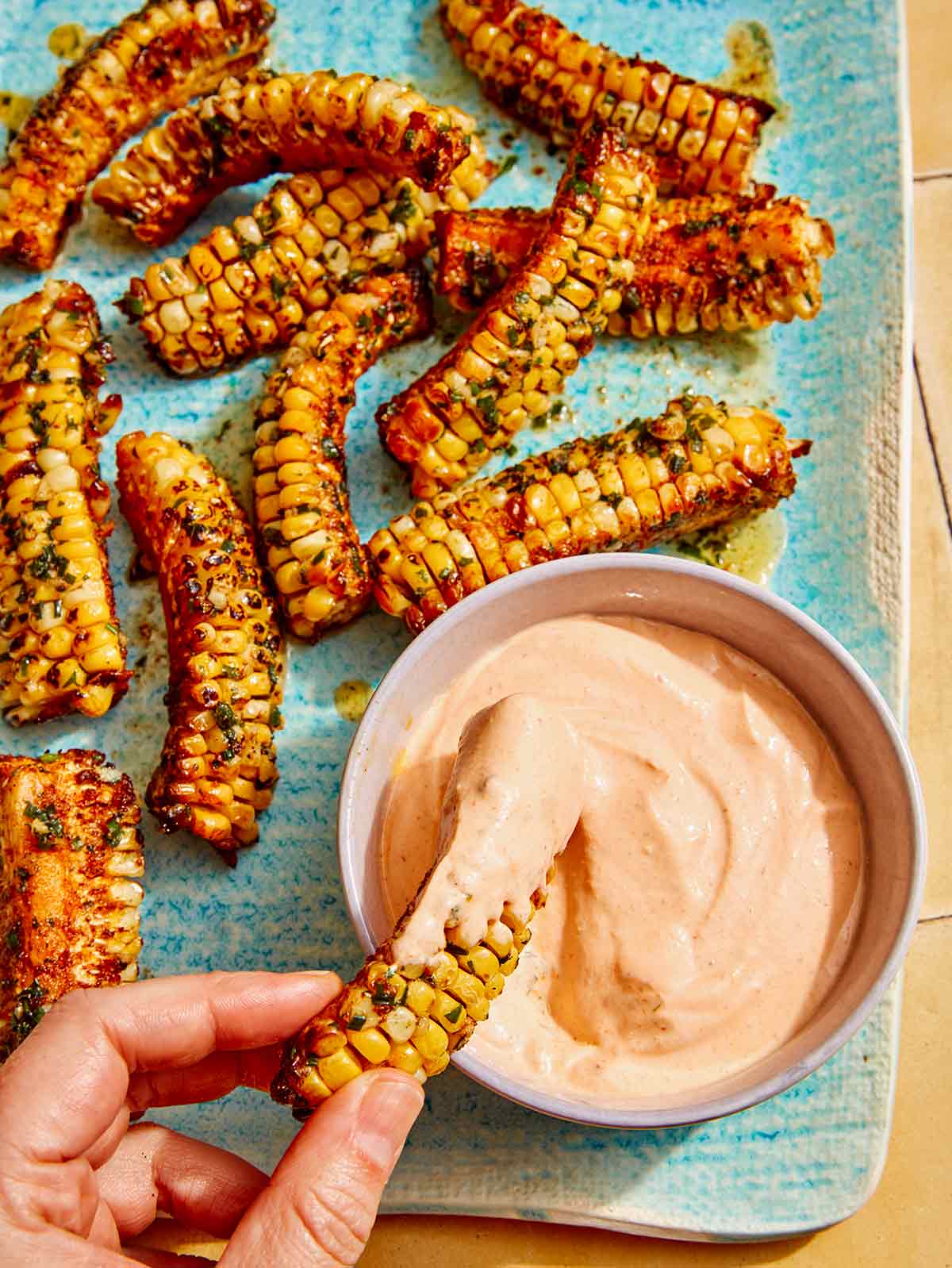 A platter of corn ribs with a dipping sauce and a hand dipping a corn rib into sauce.